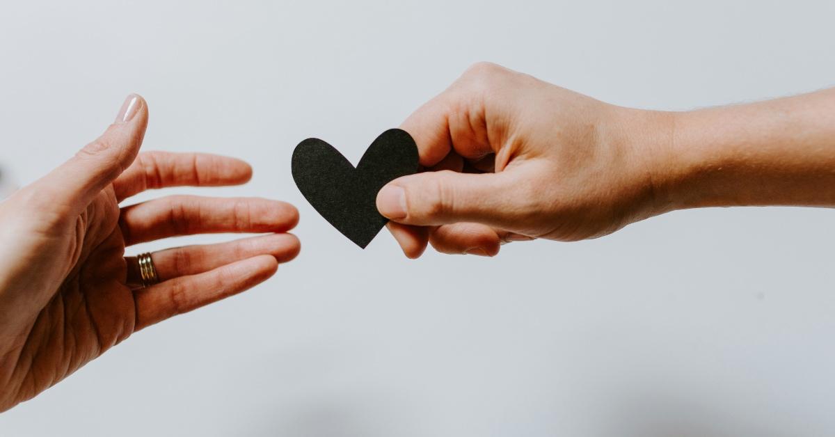 Two people with hands outstretched, one person handing a black paper heart to the other. 