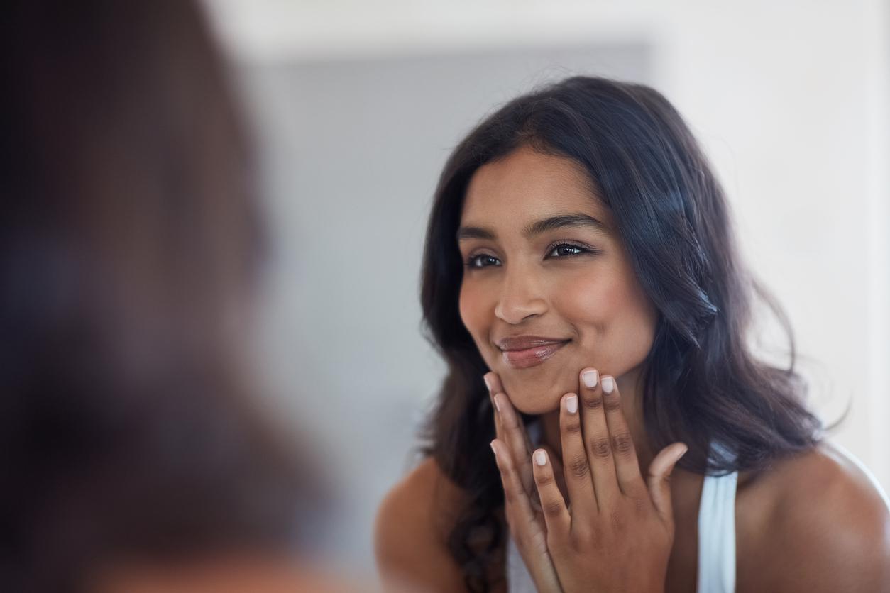 A smiling woman holds her hands to her face while looking in the mirror after practicing a healthy skin care routine.