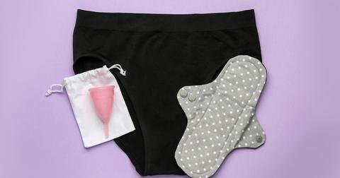 A pink menstrual cup and a gray polka-dotted reusable pad sit on top of a black pair of underwear, all on top of a purple background.