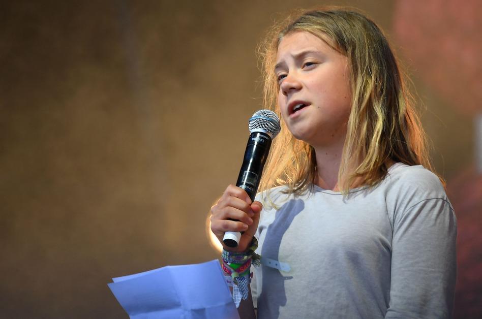 Greta Thunberg holds a microphone to her mouth and a purple piece of paper while giving a speech. 