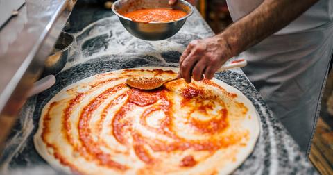 Pizza dough sits on a counter covered in flour with someone spreading tomato sauce on top.