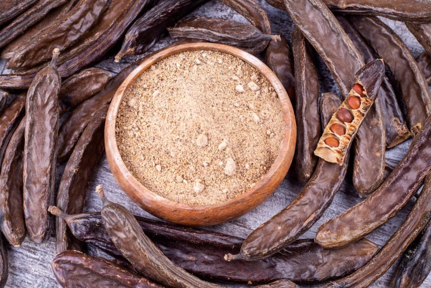 Carob pods are pictured with one exposed pod revealing carob beans, as well as ground carob in the center of the photo.