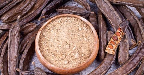 Carob pods are pictured with one exposed pod revealing carob beans, as well as ground carob in the center of the photo.