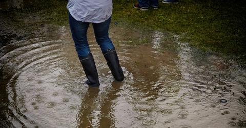 A woman stands in the middle of flood waters while wearing rainboots over her pants