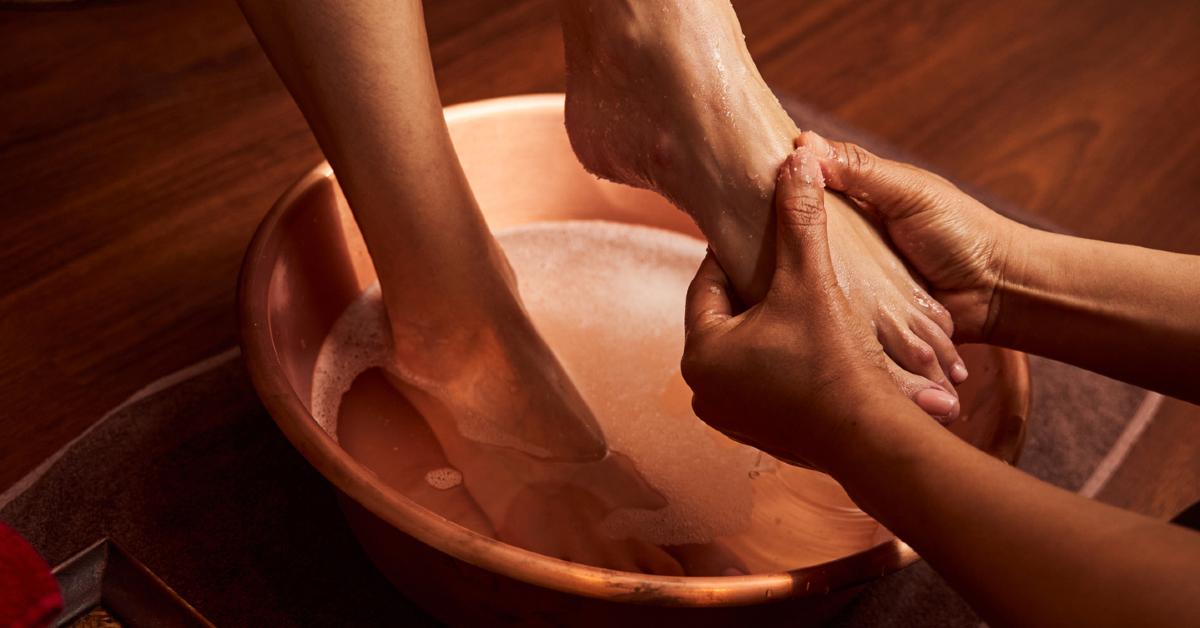 Two hands massage a woman's foot as the other soaks in a bowl of soapy water 