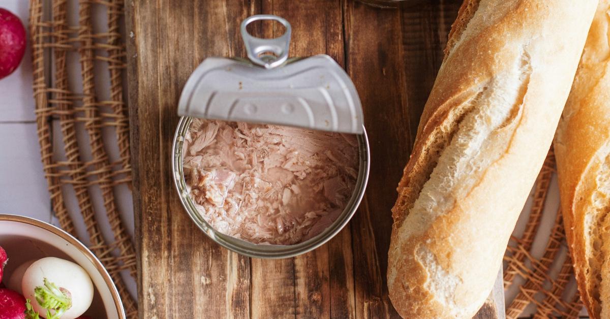 A wooden board with an opened can of canned tuna and a french baguette. 