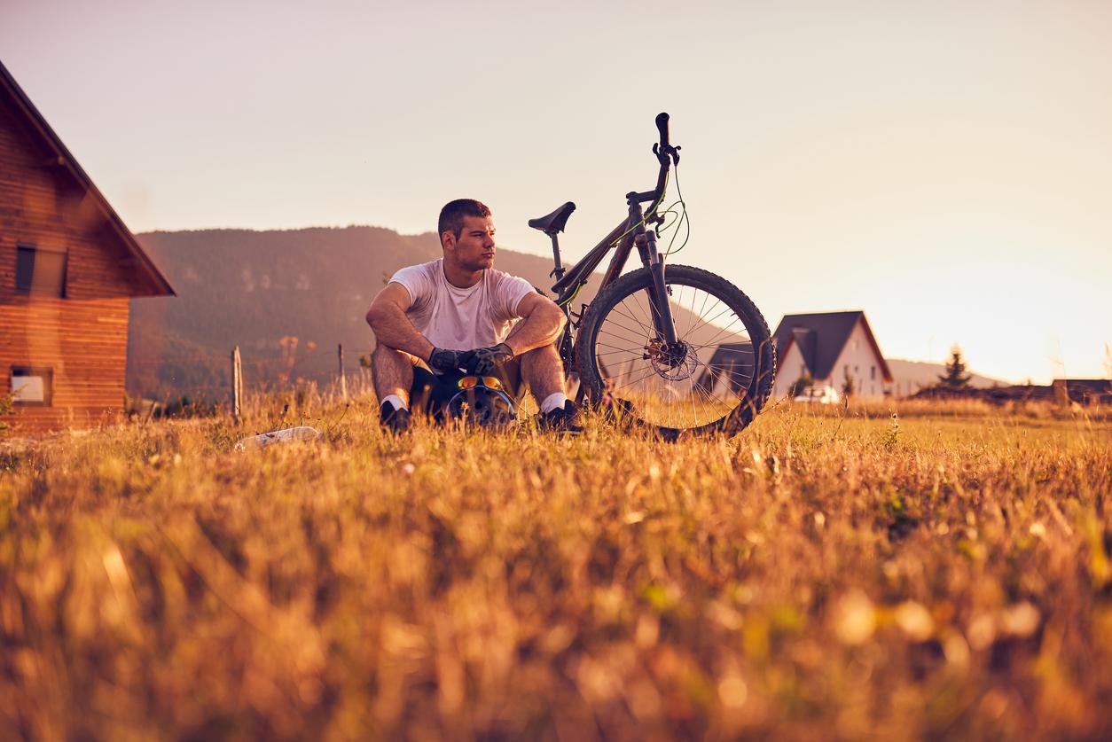 A man in a white shirt sits on a field of grass beside his bicycle.