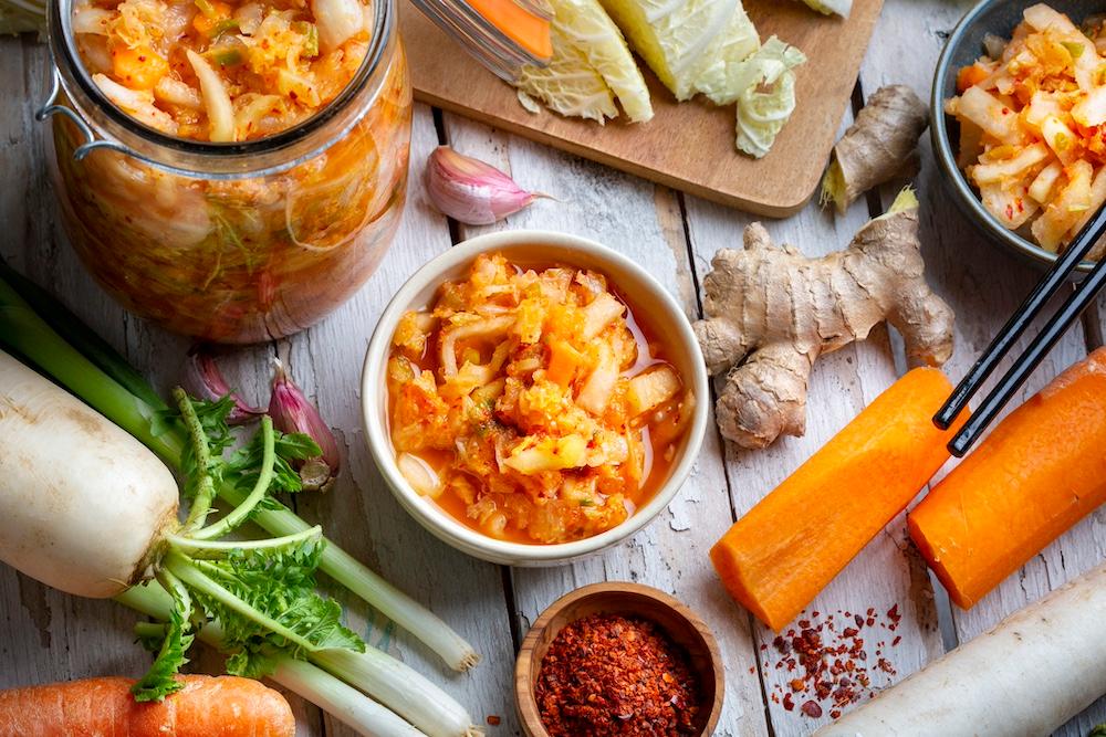 A jar and bowl of kimchi next to various ingredients on a wooden table. 