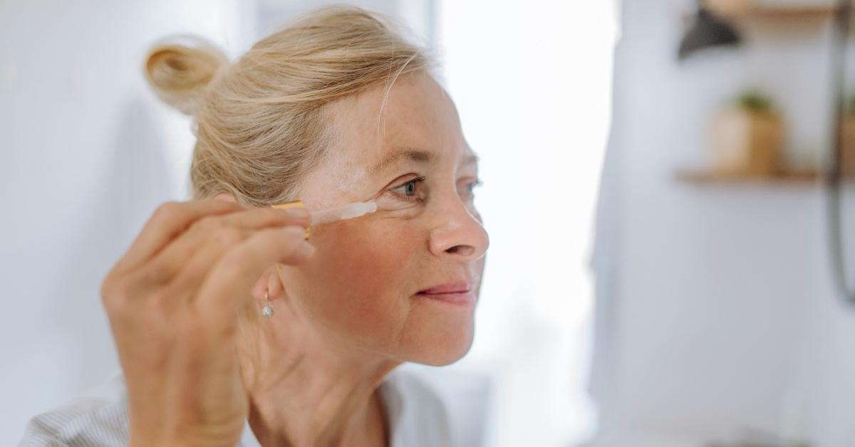 A woman looks into a mirror and applies a serum to her crow's feet.