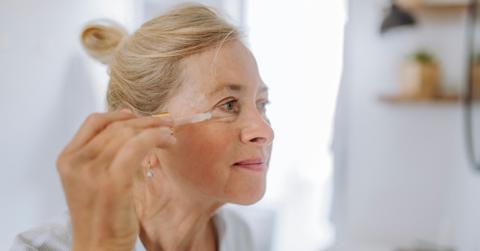 A woman looks into a mirror and applies a serum to her crow's feet.