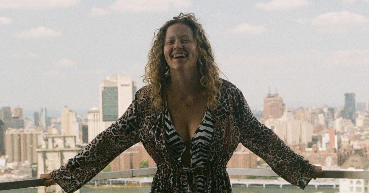 Woman with curly blonde hair standing in front of a balcony in New York City smiling. 