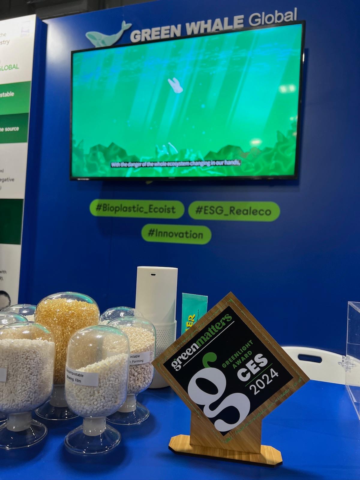 A Green Matters Greenlight Award, made of wood, sitting on a table alongside jars of pellets, in front of a TV screen and a sign reading "Green Whale Global."