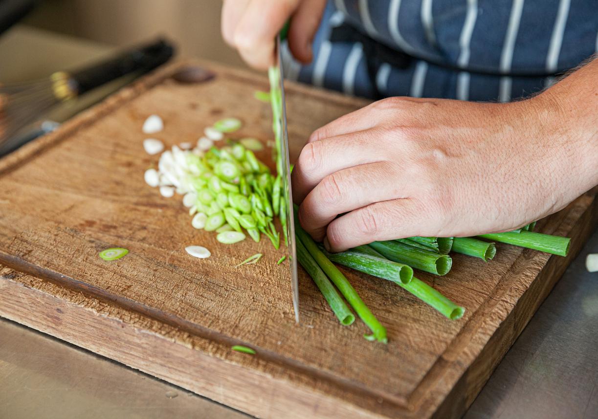 Close up of a person's hand as they chop green onions on a wood cutting board.
