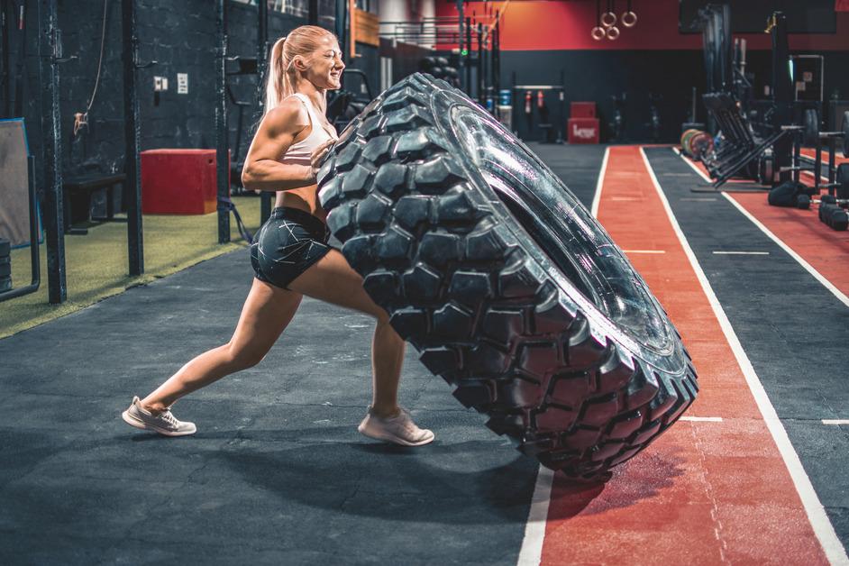 A woman with blonde hair wearing black shorts and a white shirt pushes a large tire through a CrossFit gym. 