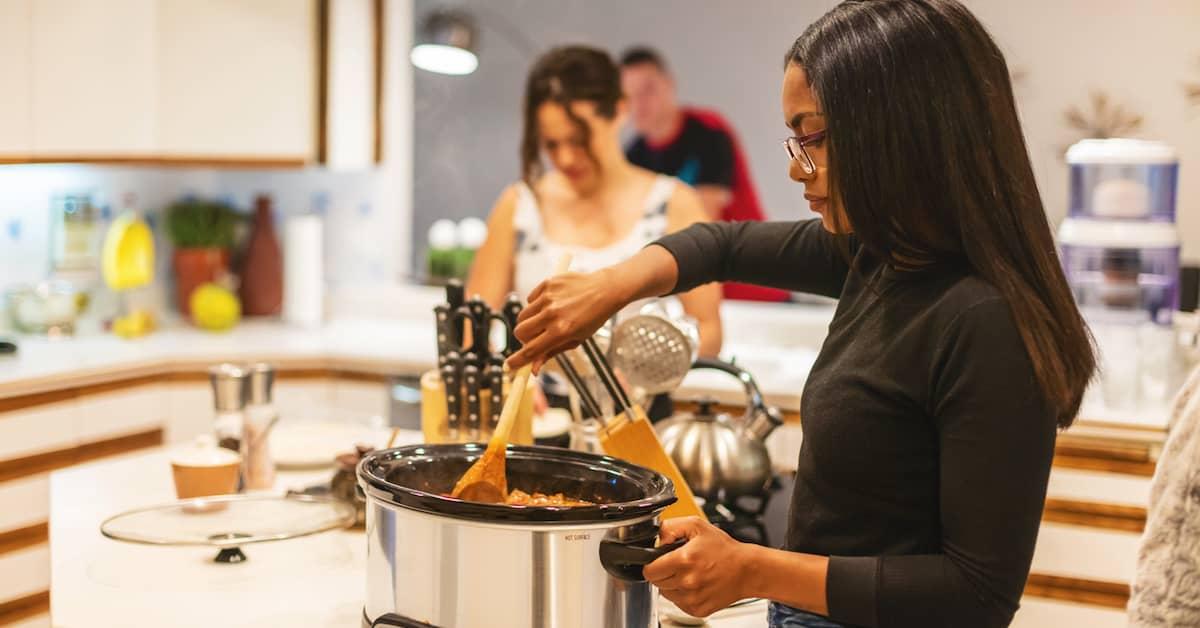 Young person in a kitchen stirring a slow cooker meal with friends in the background 