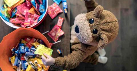 Halloween-themed photo of a child dressed up as a teddy bear standing in front of a basket of candy bars (Representative Cover Image Source: Getty Images | Onfokus)