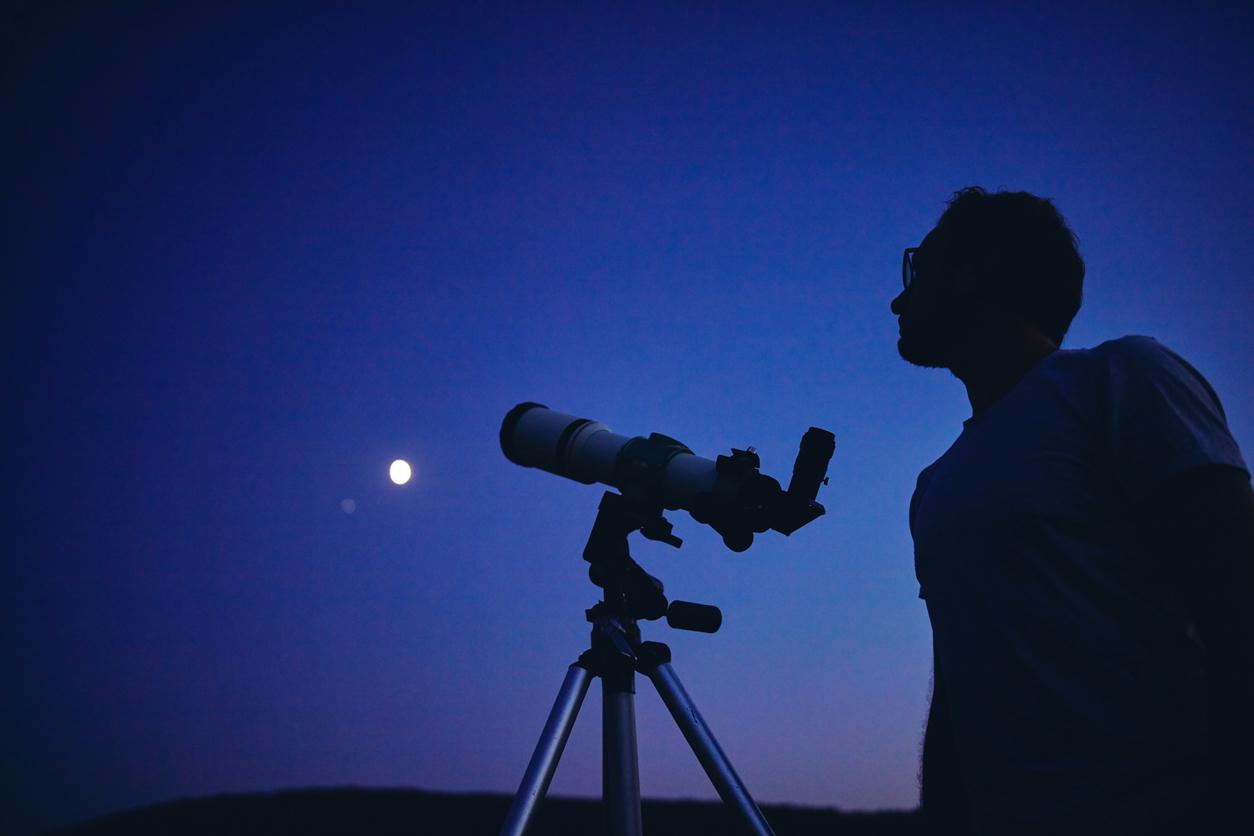 A man looks up towards the night sky beside a telescope.