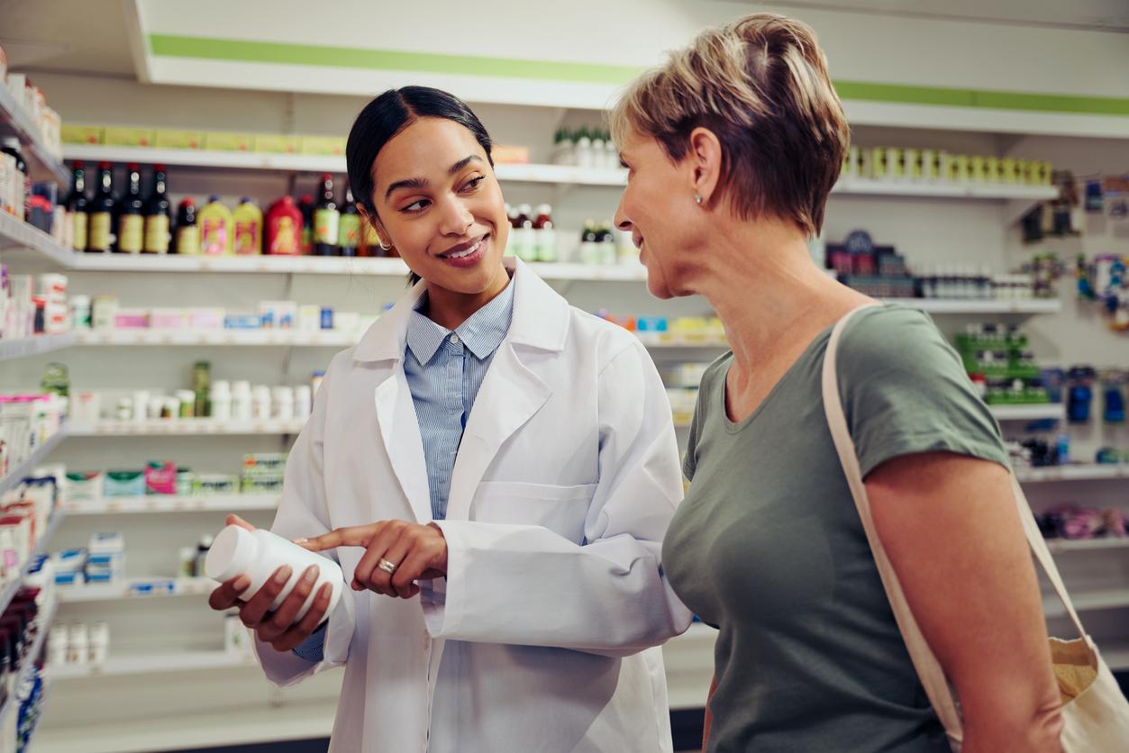 A female pharmacist happily explains a medication to a female customer while pointing to a white pill bottle in a pharmacy.