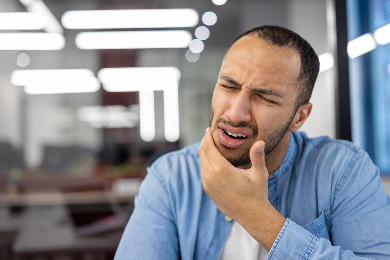 A man at an office winces in pain while placing his hand on his cheek.