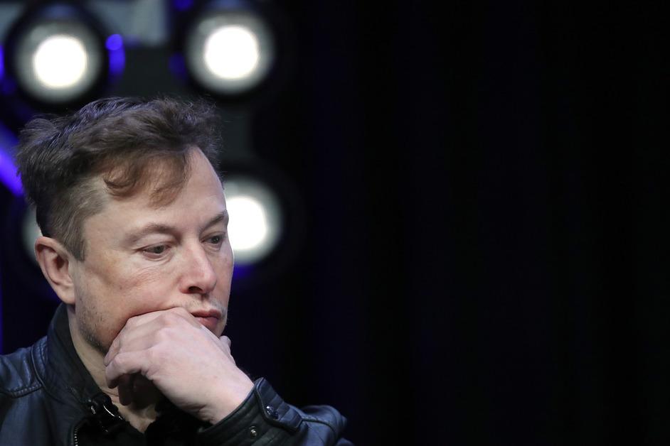 A close-up photo of Elon Musk's face with his hand covering his mouth and a perplexed expression. 