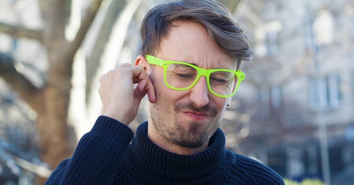 Young person with green neon glasses on, holding their hand up to their right ear and grimacing in pain