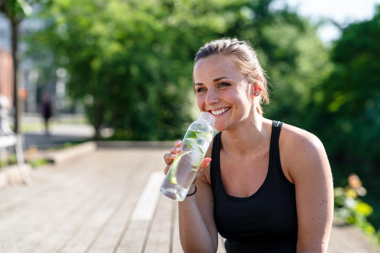 A smiling woman in a black tank top consumes a bottle with cucumber water.