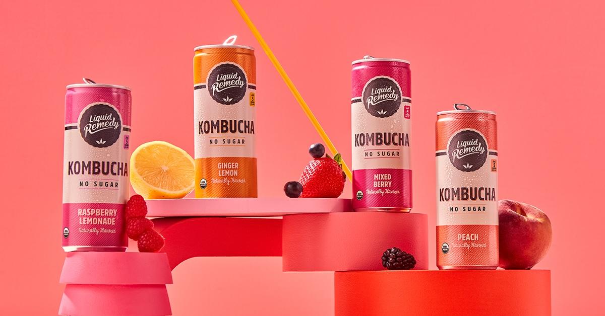 Four Liquid Remedy kombucha cans alongside pieces of fruit on an orange and pink display.