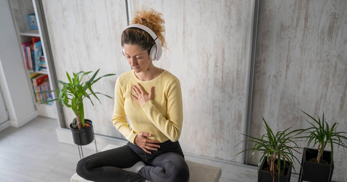 Young person sitting cross legged in a yoga position, eyes closed, hands placed on chest and belly with earphones on.