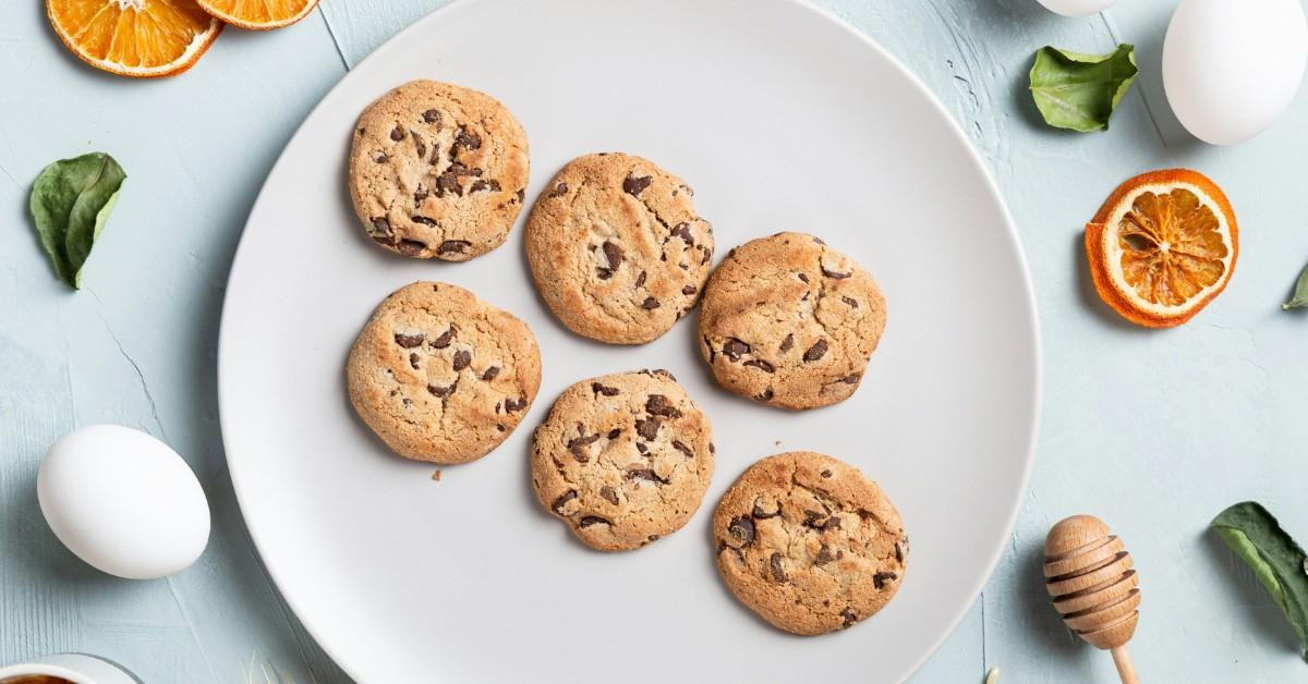 Fresh baked chocolate chip cookies sit on a plate amongst several other key ingredients 