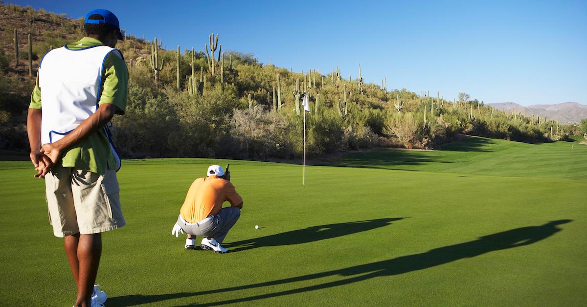 Two men playing golf on a course in Arizona, one man in an orange shirt crouches down by the ball.