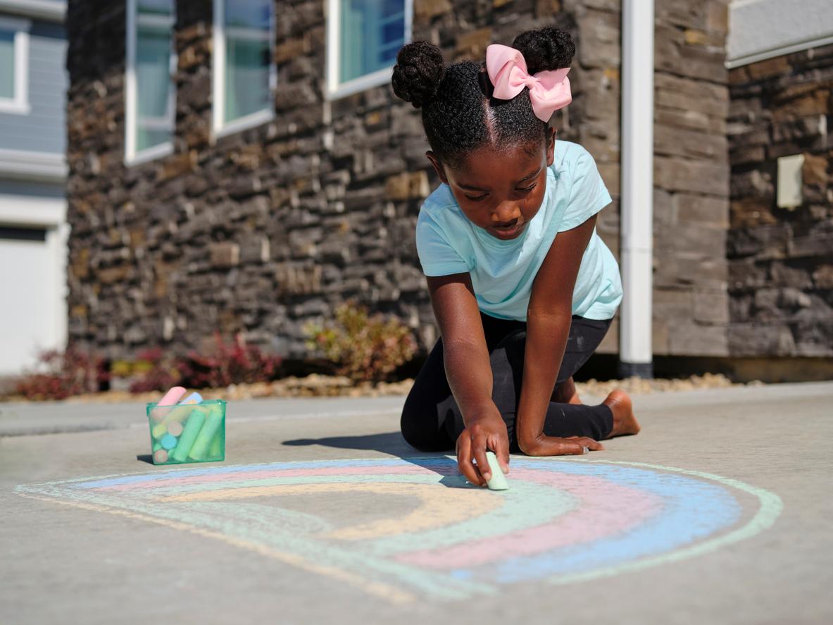 A young child plays with colorful chalk on a sidewalk, using a green-colored piece of chalk to color in a rainbow.