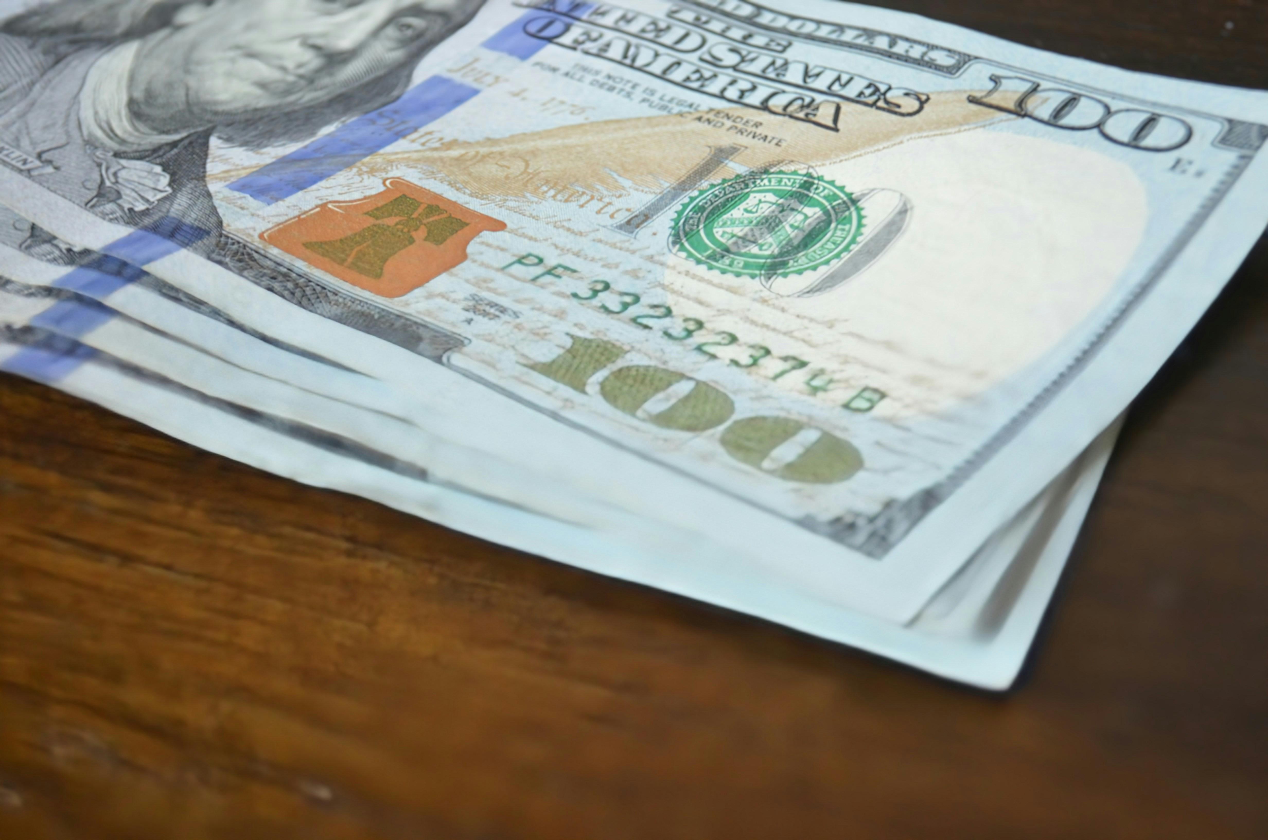 A stack of four 100-dollar bills lays atop a wooden table.