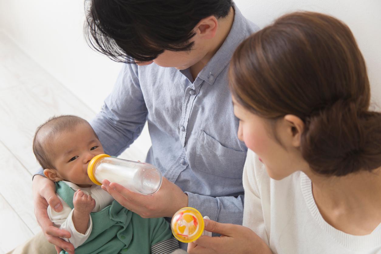 A couple feeds their baby through a glass milk bottle while the husband holds the baby in his arms.