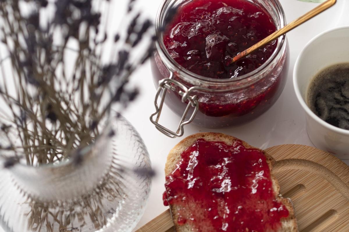 Jar of raspberry jam next to toast with jam and a vase.