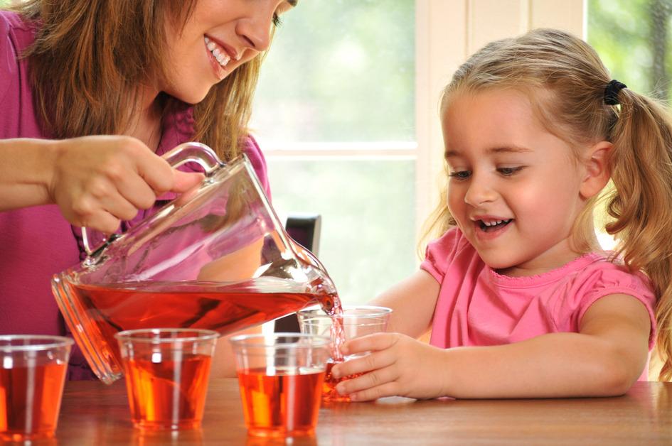 A woman wearing a pink shirt pours a red drink into a glass held by a girl in pigtails wearing a light pink shirt. 