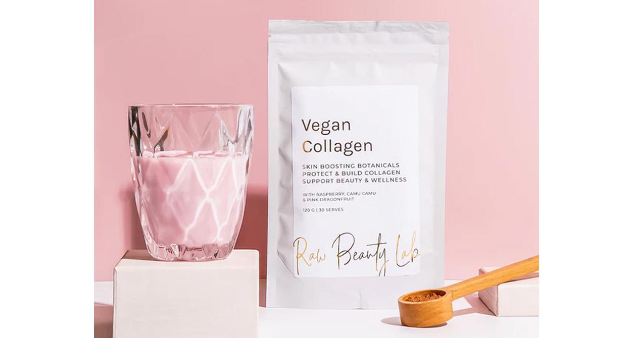 Package of Raw Beauty Lab Vegan Collagen Superfood sits next to a glass of a pink liquid and a wooden spoon, in front of a pink background.