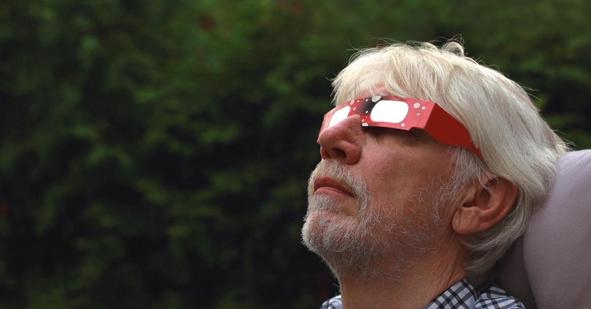 An older man sits in a chair while wearing red solar eclipse glasses and looking at the sky.