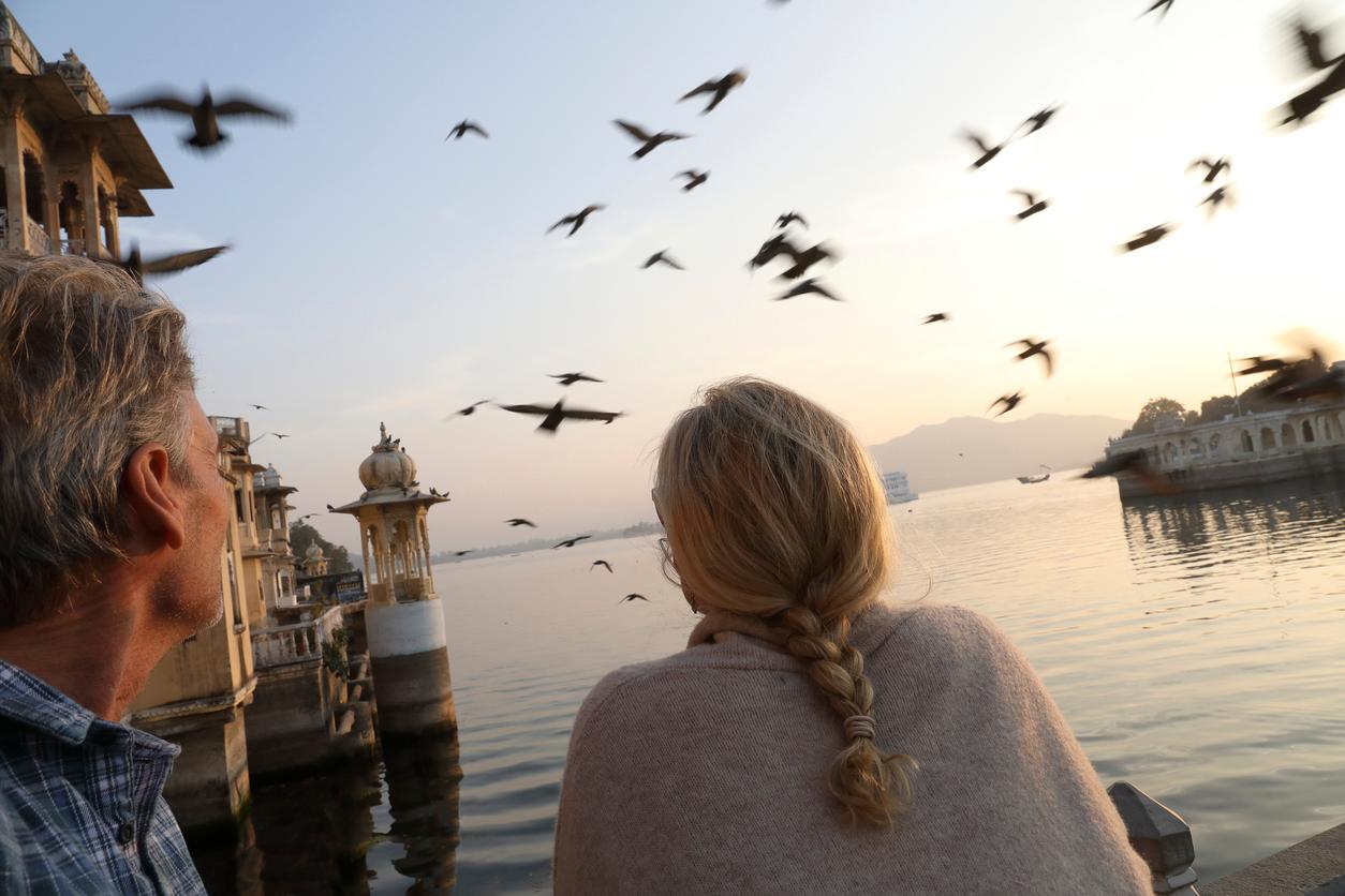 A man and woman smile as they look out at a flock of birds flying above a body of water.