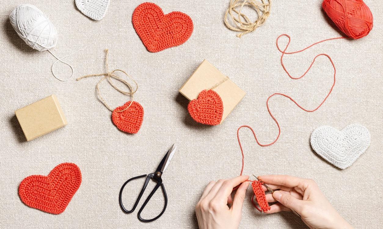 View of someone's hands while working on a crochet project near red and white crochet hearts and yarn.