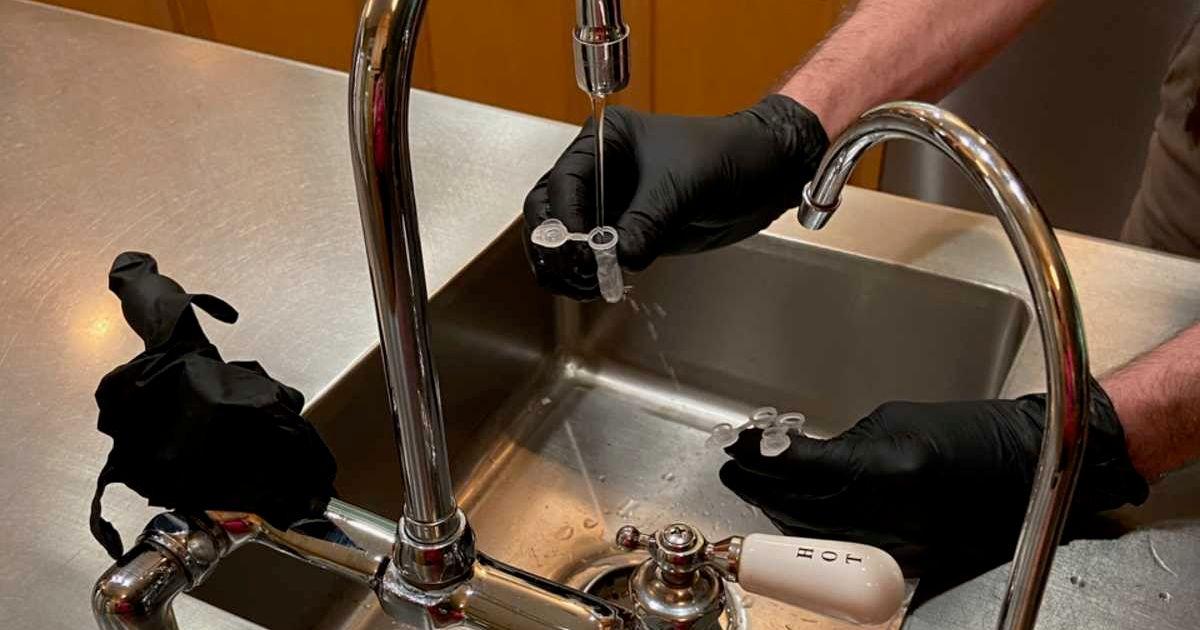 A USGS scientist wearing black gloves is collecting a sample of tap water from the kitchen sink using small plastic vials to test for PFAS. (Image Source: USGS)
