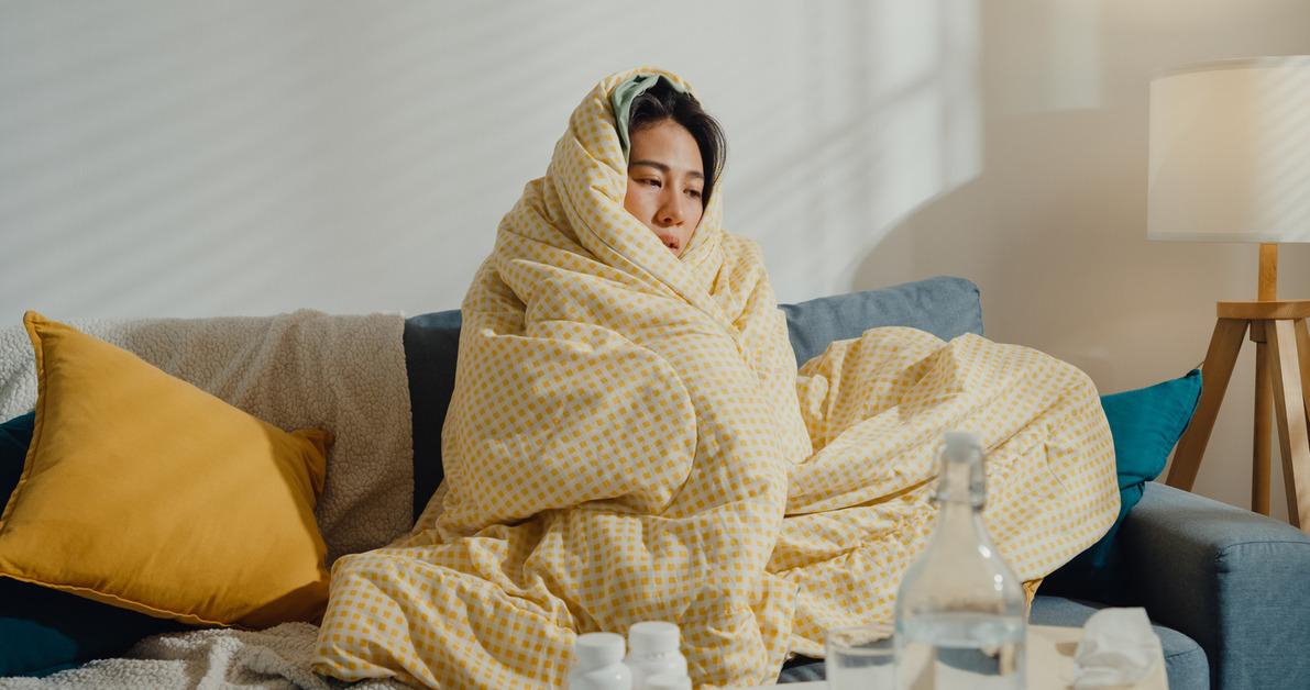 A woman sits on the couch wrapped in a yellow blanket while looking ill. 