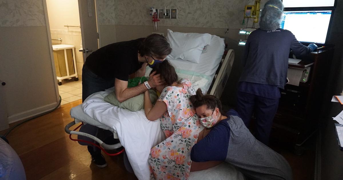 A woman in labor kneels near a hospital bed, with two people hugging her.