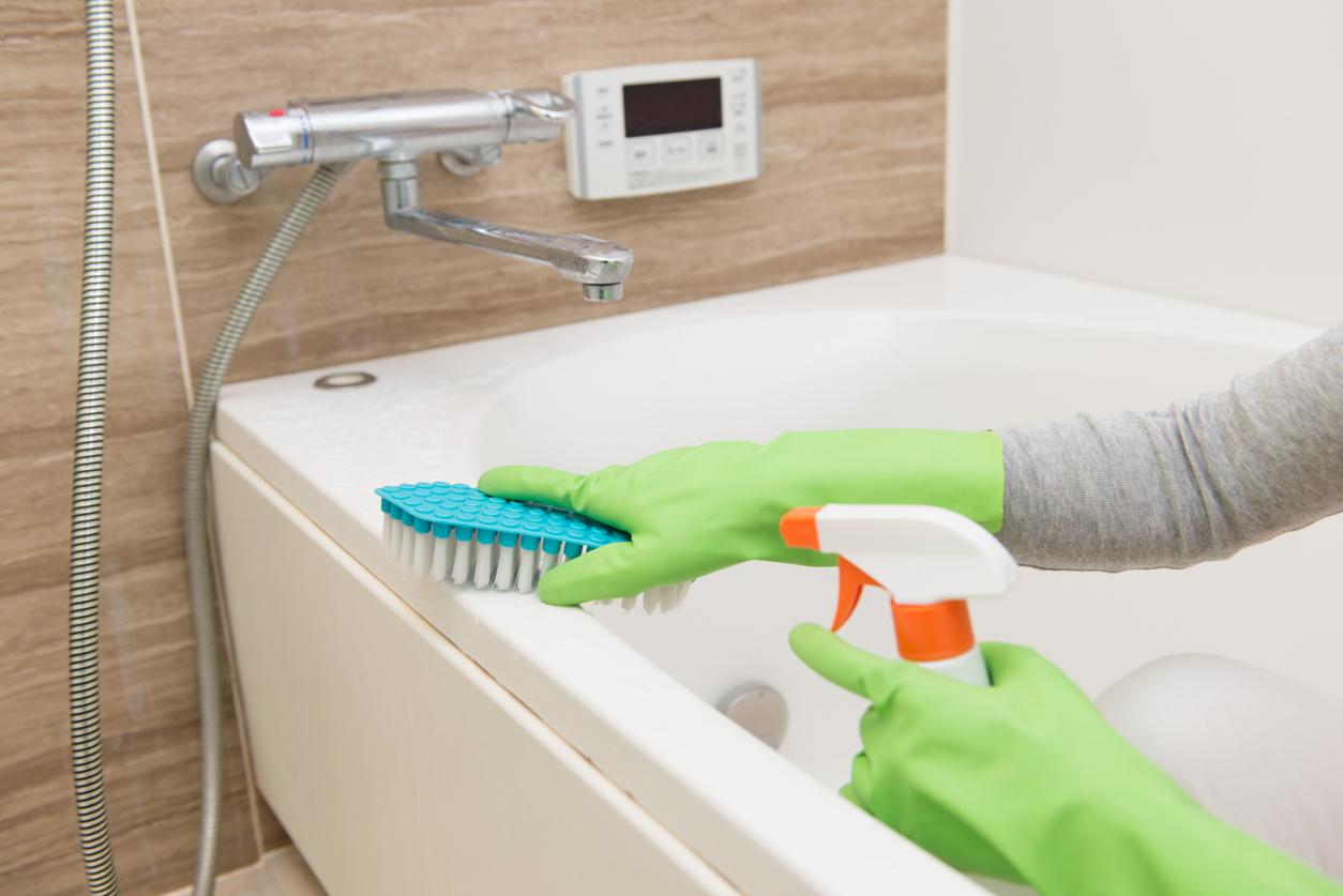 A person wearing green rubber cleaning gloves and scrubbing the rim of a bathtub with a brush.