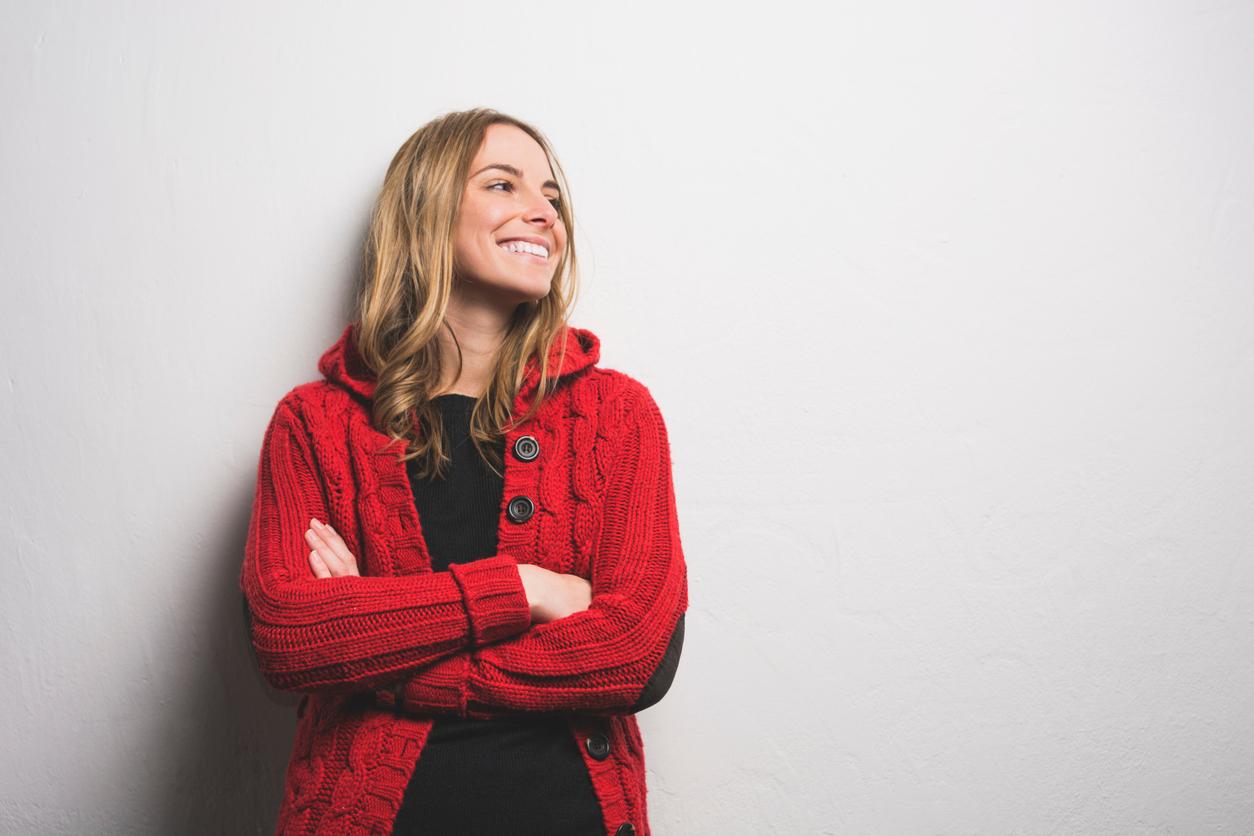 Person with long, blonde hair leaning against a white wall wearing a bright red button-up cardigan over a black top.