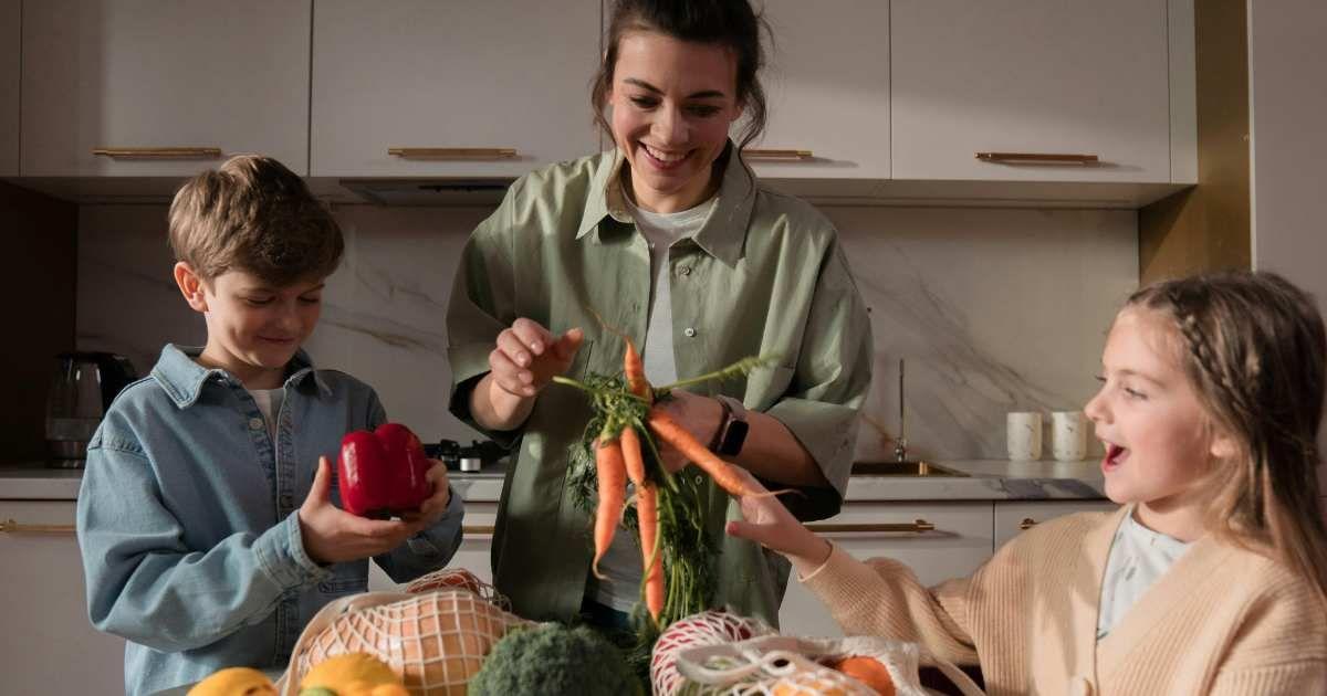A mom holding a bunch of fresh orange carrots and her children smiling next to her in the kitchen. (Representative Cover Image Source: Pexels | Ron Lach)