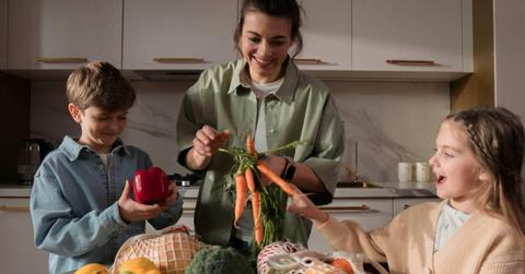 A mom holding a bunch of fresh orange carrots and her children smiling next to her in the kitchen. (Representative Cover Image Source: Pexels | Ron Lach)