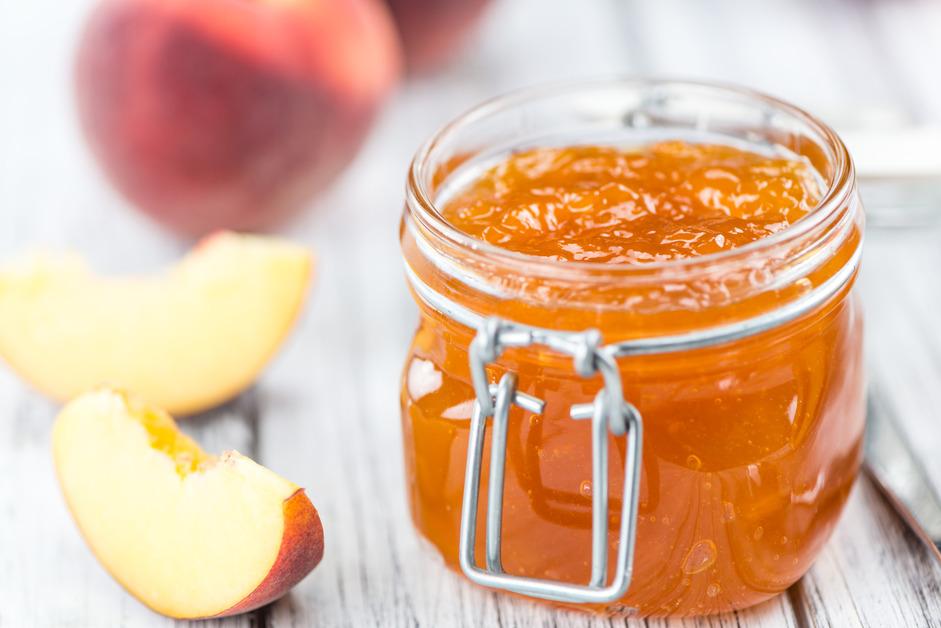 A jar of peach jam open next to a couple slices of peaches. 