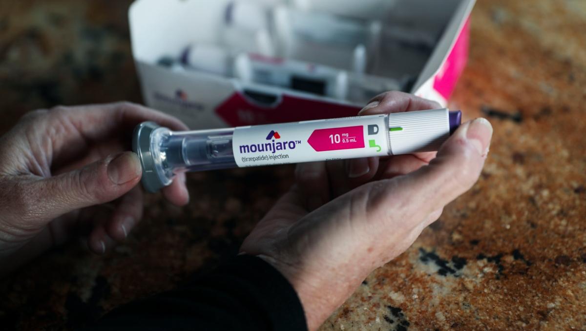 A person holds up a vial of Mounjaro medication in front of the box.