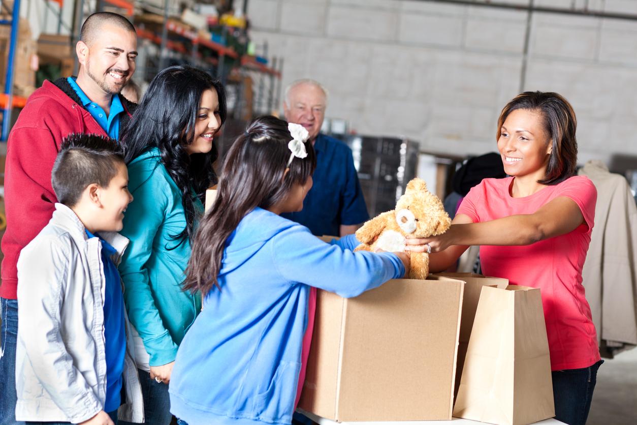 A smiling volunteer gives a girl a teddy bear with her smiling family beside her.