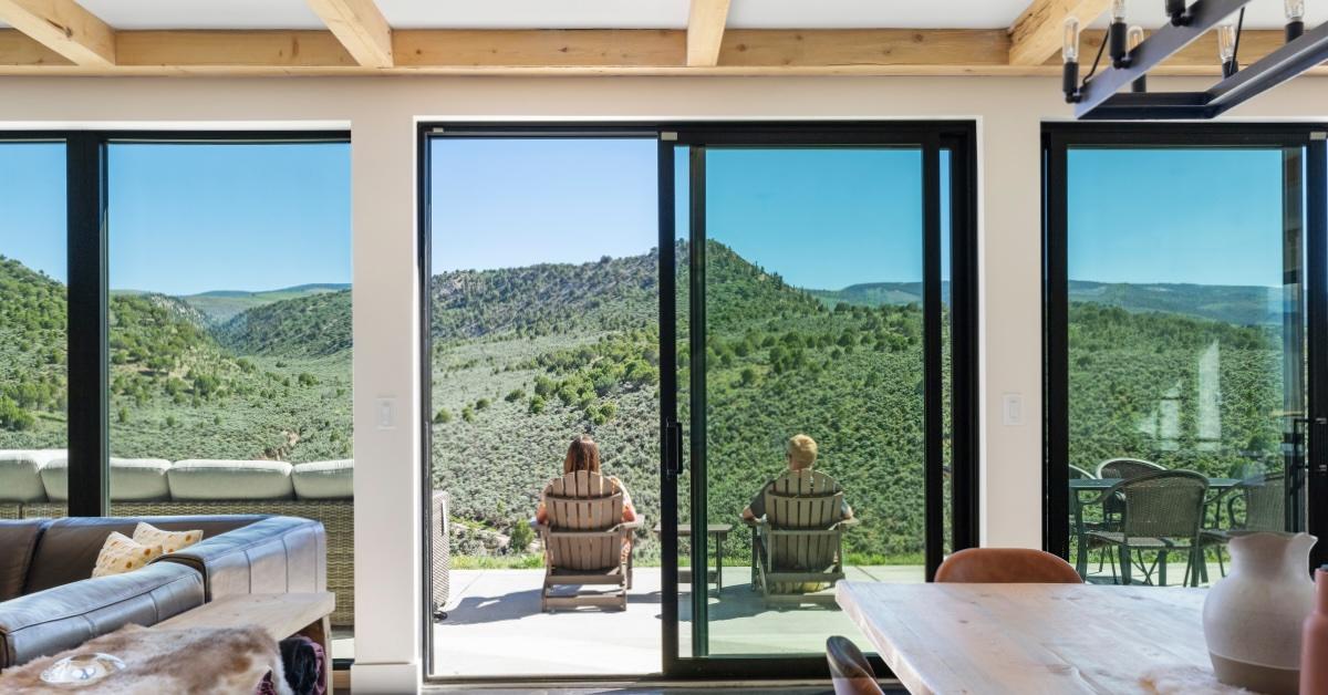 Two people sit in Adirondack chairs on a deck overlooking lush mountains.